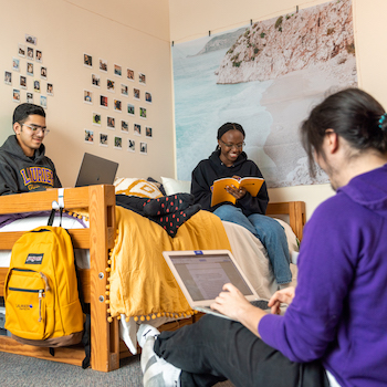 three students in residence room sitting on bed studying